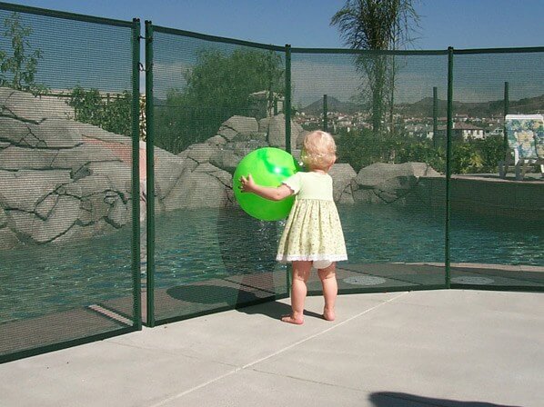 toddler holding a green ball leaning against a mesh pool safety fence