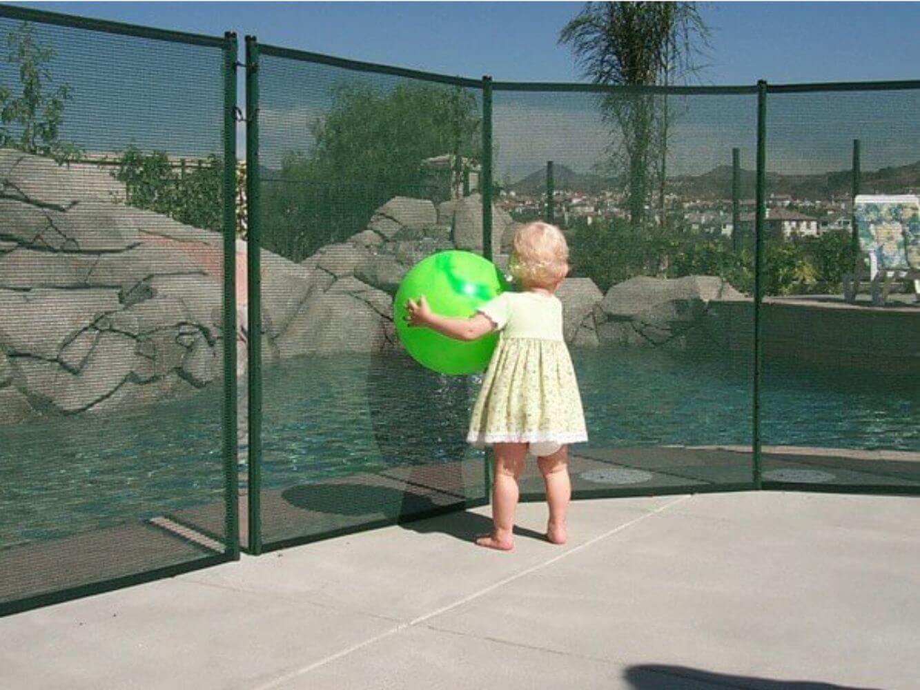 Toddler holding a large ball standing in front of a pool safety fence