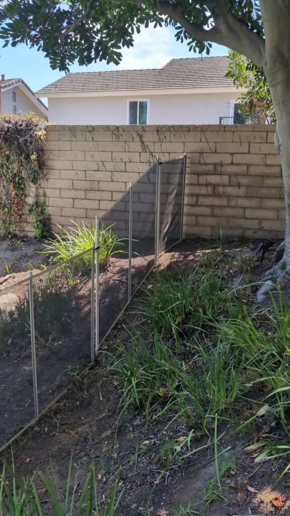 Beige mesh pool fence stepping down a sloped planter bed beside a block wall