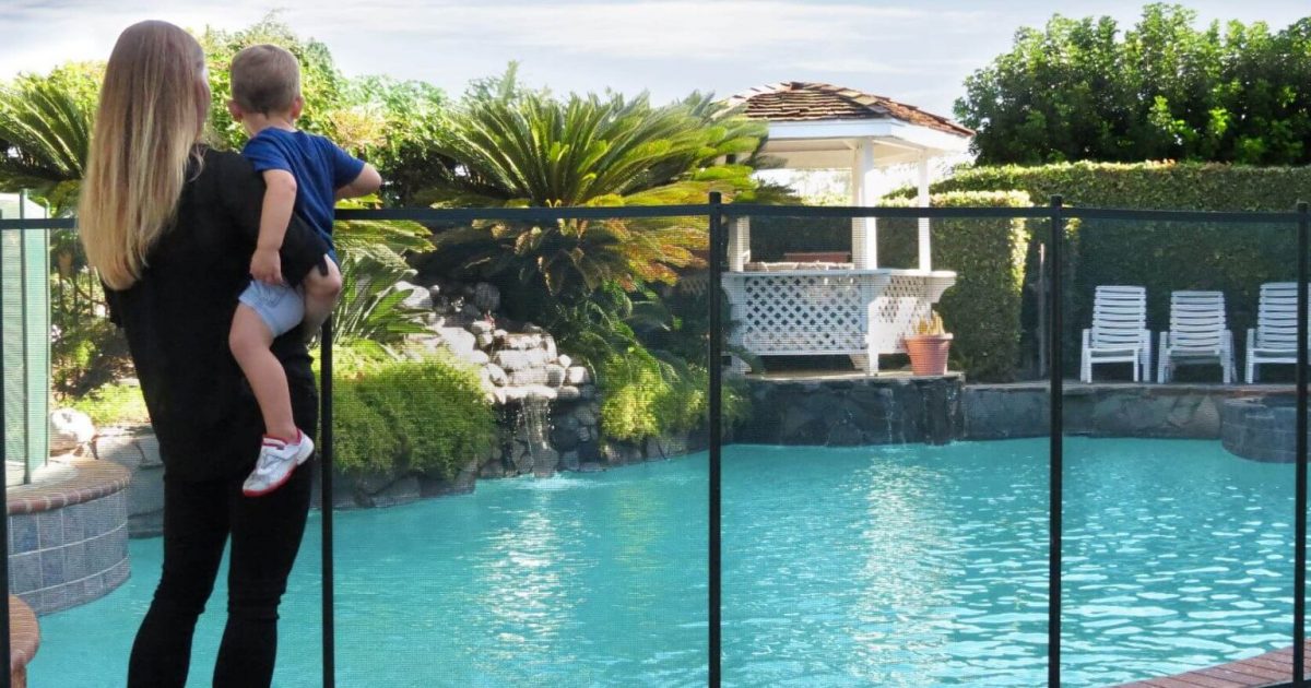 Mother holding her toddler while they stand in front of an installed mesh pool fence around the swimming pool